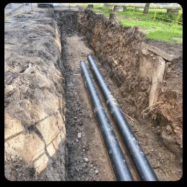 Two black pipes sit at the bottom of a deep trench dug into the earth