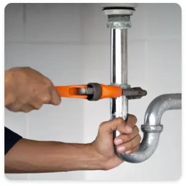 A person repairs a leaky pipe with a pipe wrench under a sink
