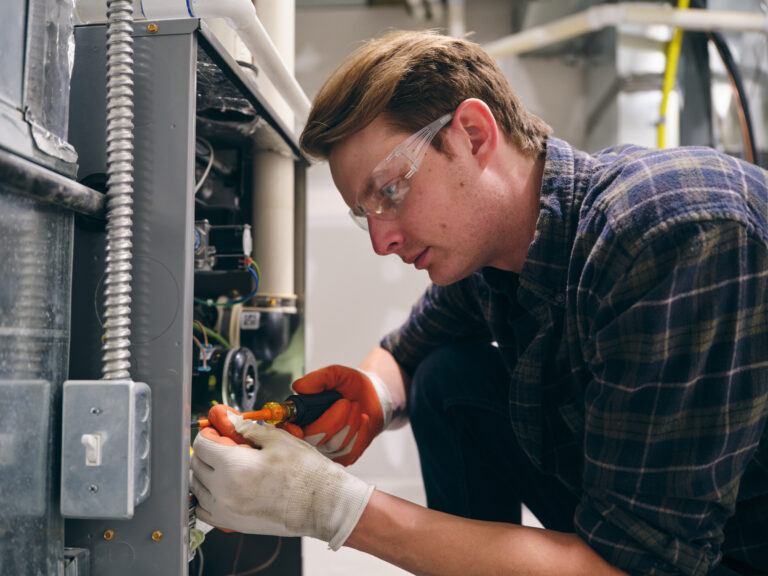 A technician wearing safety goggles and gloves is focused on working on electrical components inside a machine, using a screwdriver to tighten a connection. The background features pipes and electrical wiring.