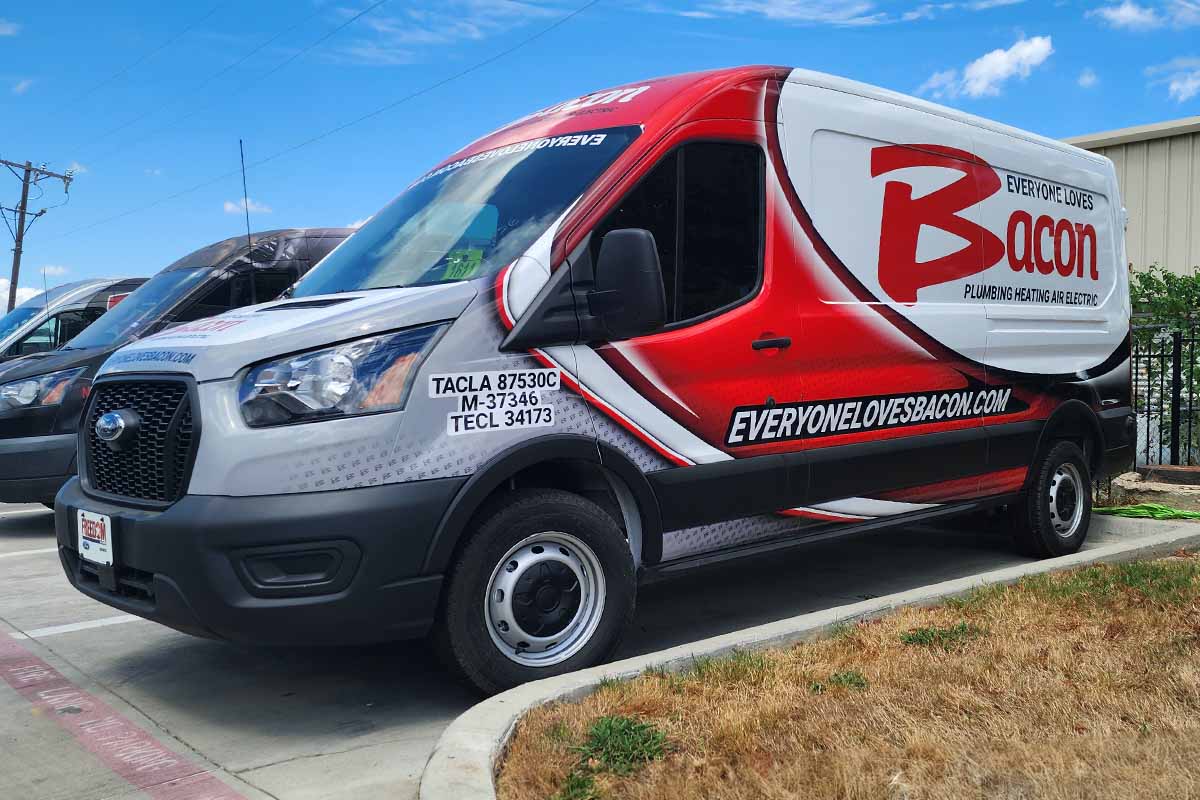 A Bacon Plumbing Heating Air Electric service van with "Everyone Loves Bacon" prominently displayed on the side is parked in a lot. The van's design features red, black, and white colors along with license and certification numbers near the driver's door.