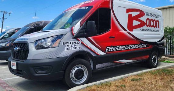 A Bacon Plumbing Heating Air Electric service van with "Everyone Loves Bacon" prominently displayed on the side is parked in a lot. The van's design features red, black, and white colors along with license and certification numbers near the driver's door.