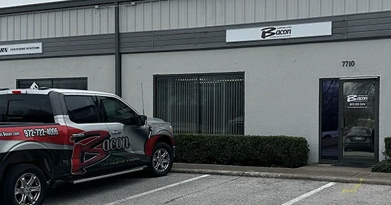 Bacon Plumbing Heating Air Electric office in Fort Worth, TX, featuring a gray commercial building with the company’s signage above the entrance and on the door. A branded Bacon service truck is parked in front of the office.